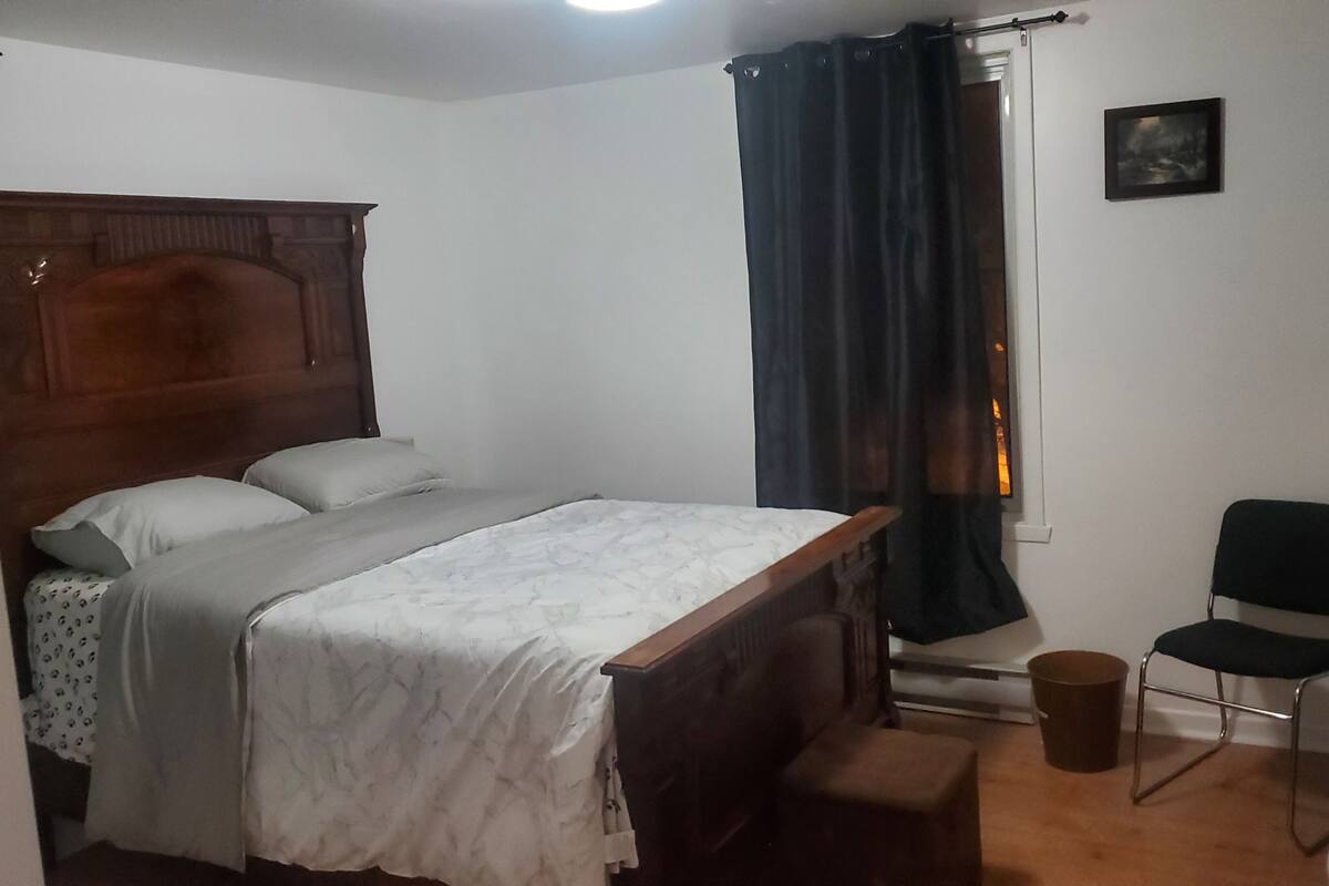 Une chambre à coucher avec un lit en bois, une chaise, une petite table et une fenêtre avec des rideaux.