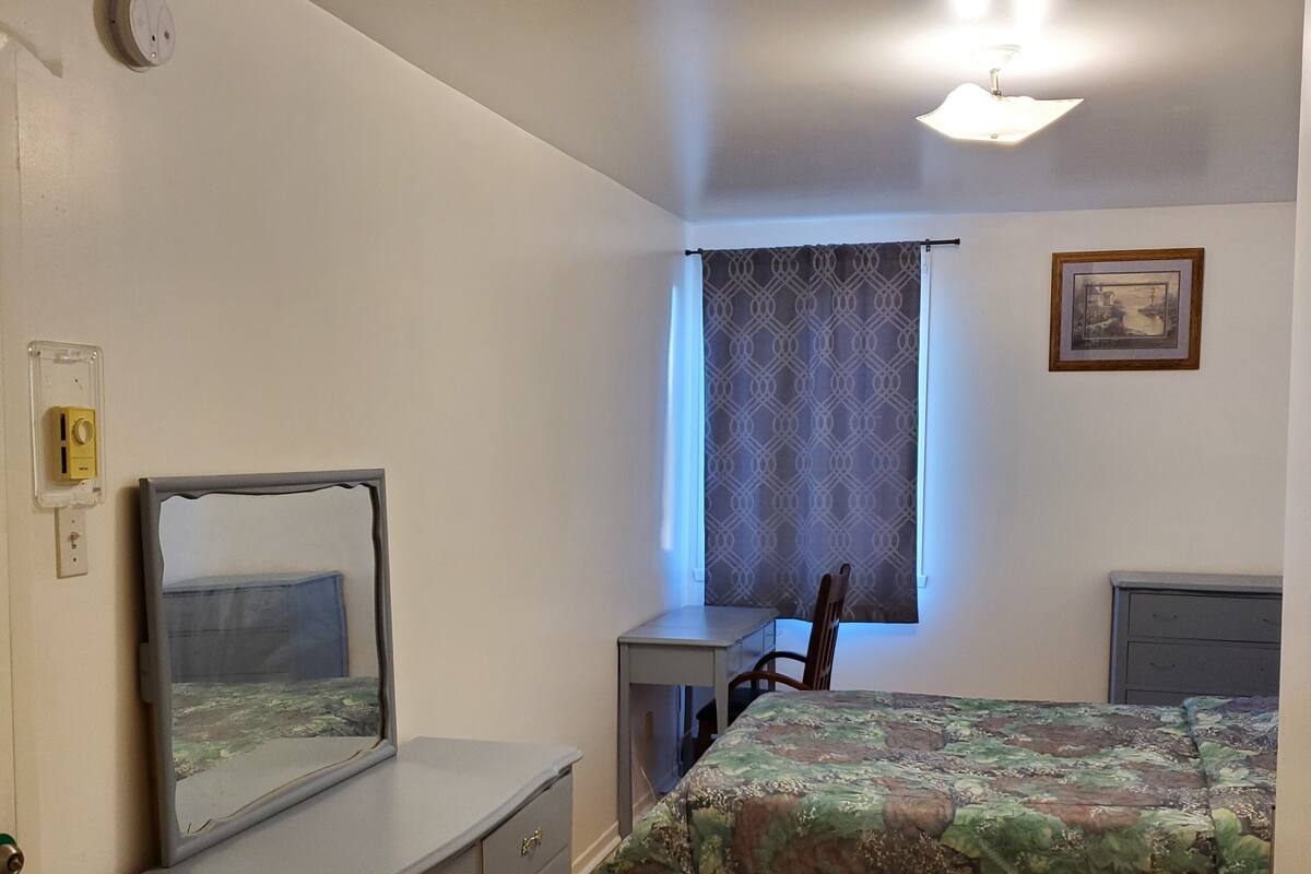 Une chambre à coucher comprenant un lit, une commode, un bureau, une chaise, une fenêtre avec des rideaux, un luminaire fixé au mur et un tableau encadré.