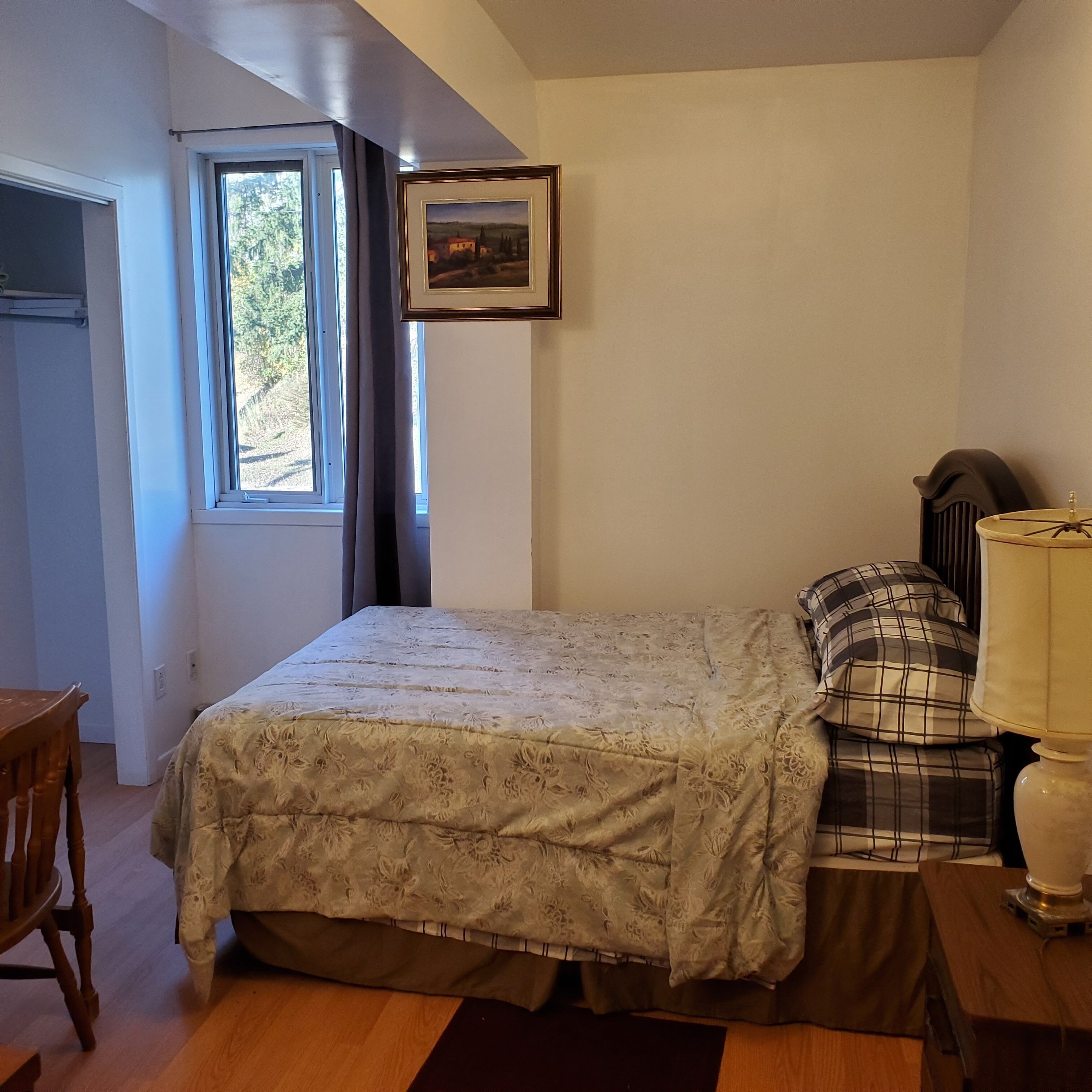 Un dormitorio con cama, ventana, un cuadro en la pared, lámpara y una silla.