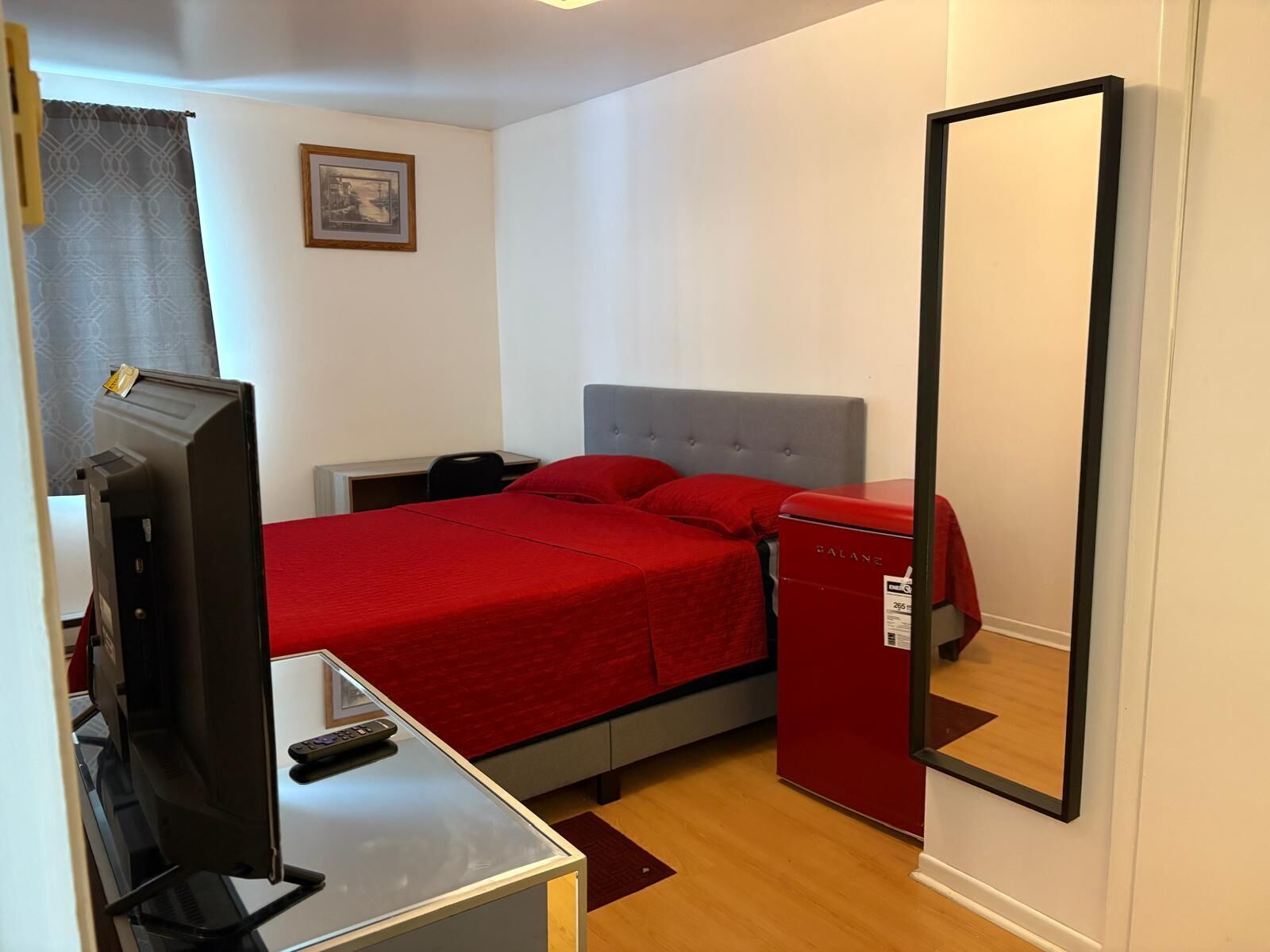 A bedroom with a bed, a television, a small refrigerator, and a mirror.
