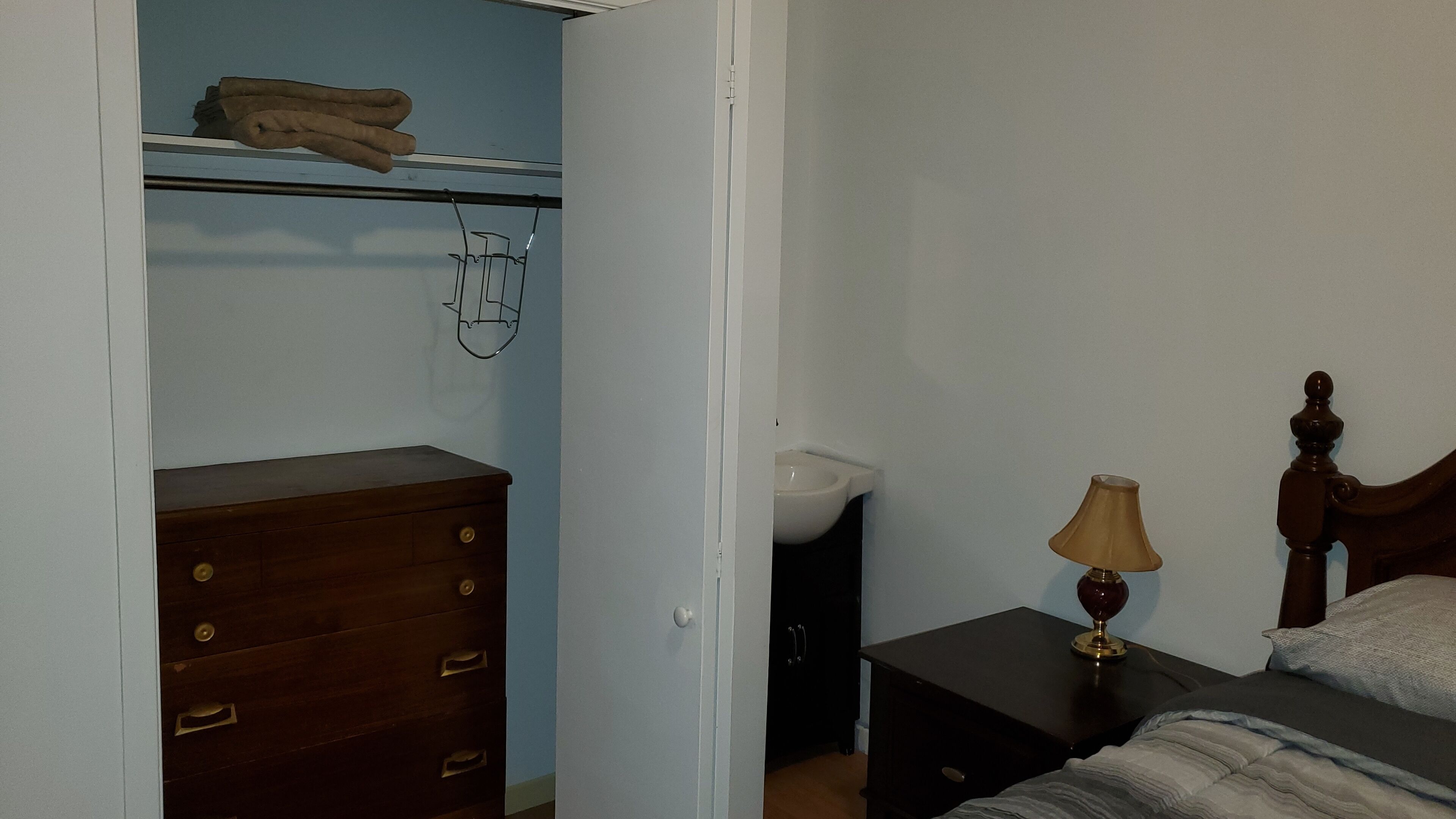 A bedroom with a wooden dresser, a bed with a wooden headboard, a bedside table with a lamp, and a closet with a towel rack.