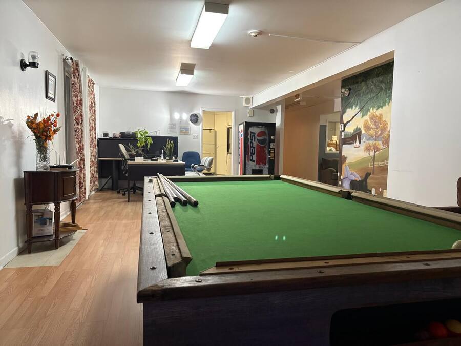 Une salle de billard équipée d’une table de billard, d’un bureau avec un ordinateur et d’une grande fenêtre donnant sur un parc.