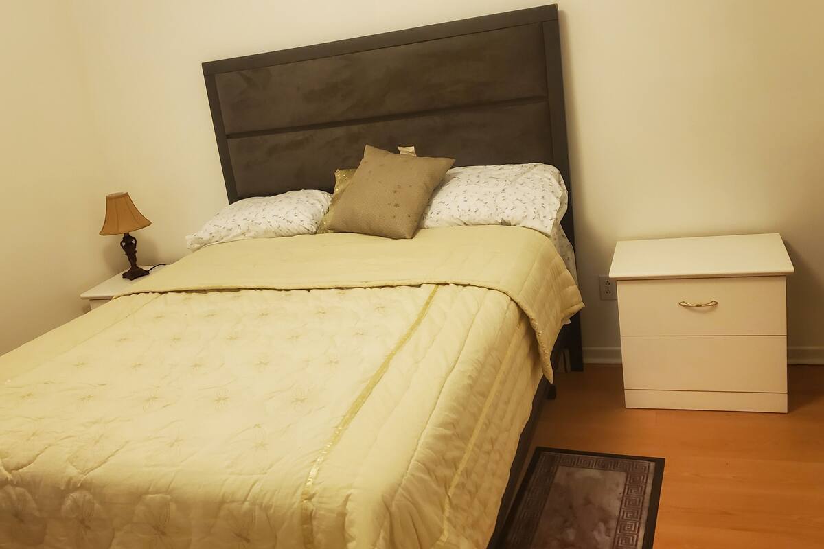 Un lit bien fait, avec une tête de lit, une table de chevet dotée d’une lampe et un petit tiroir sur le côté.