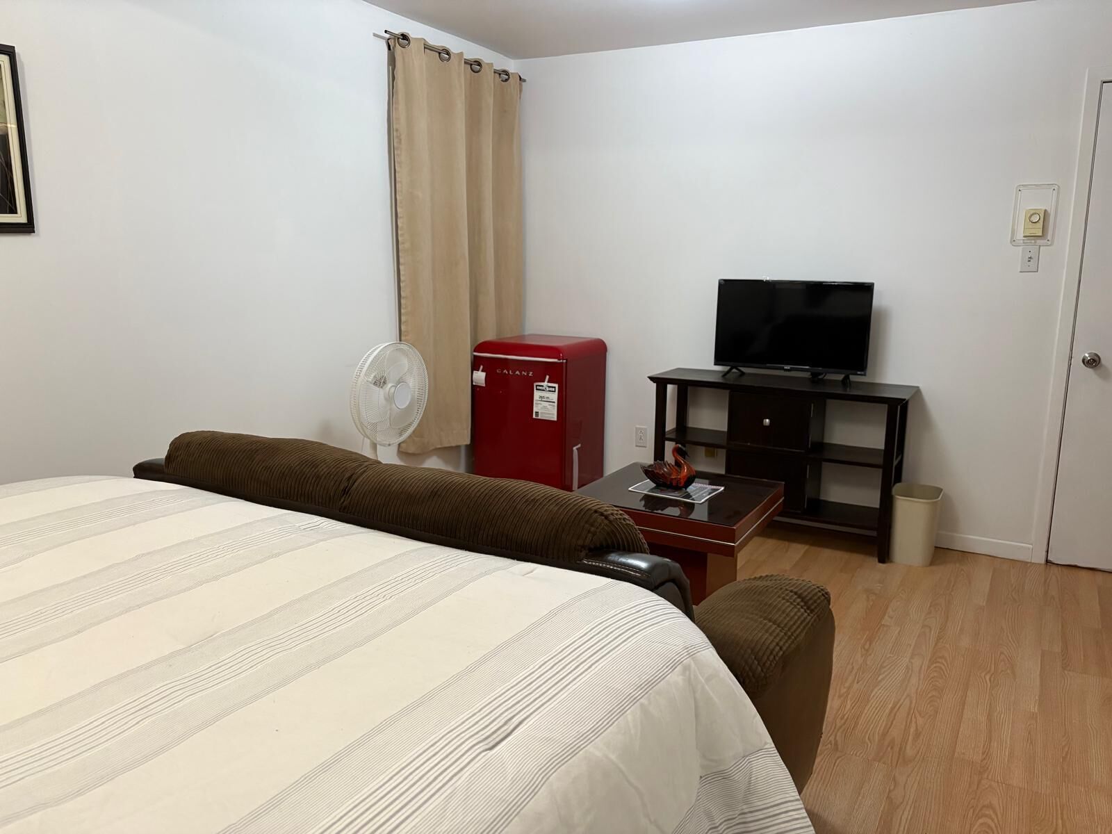 A hotel room with a bed, a red refrigerator, a television on a stand, a fan, and a wooden floor.
