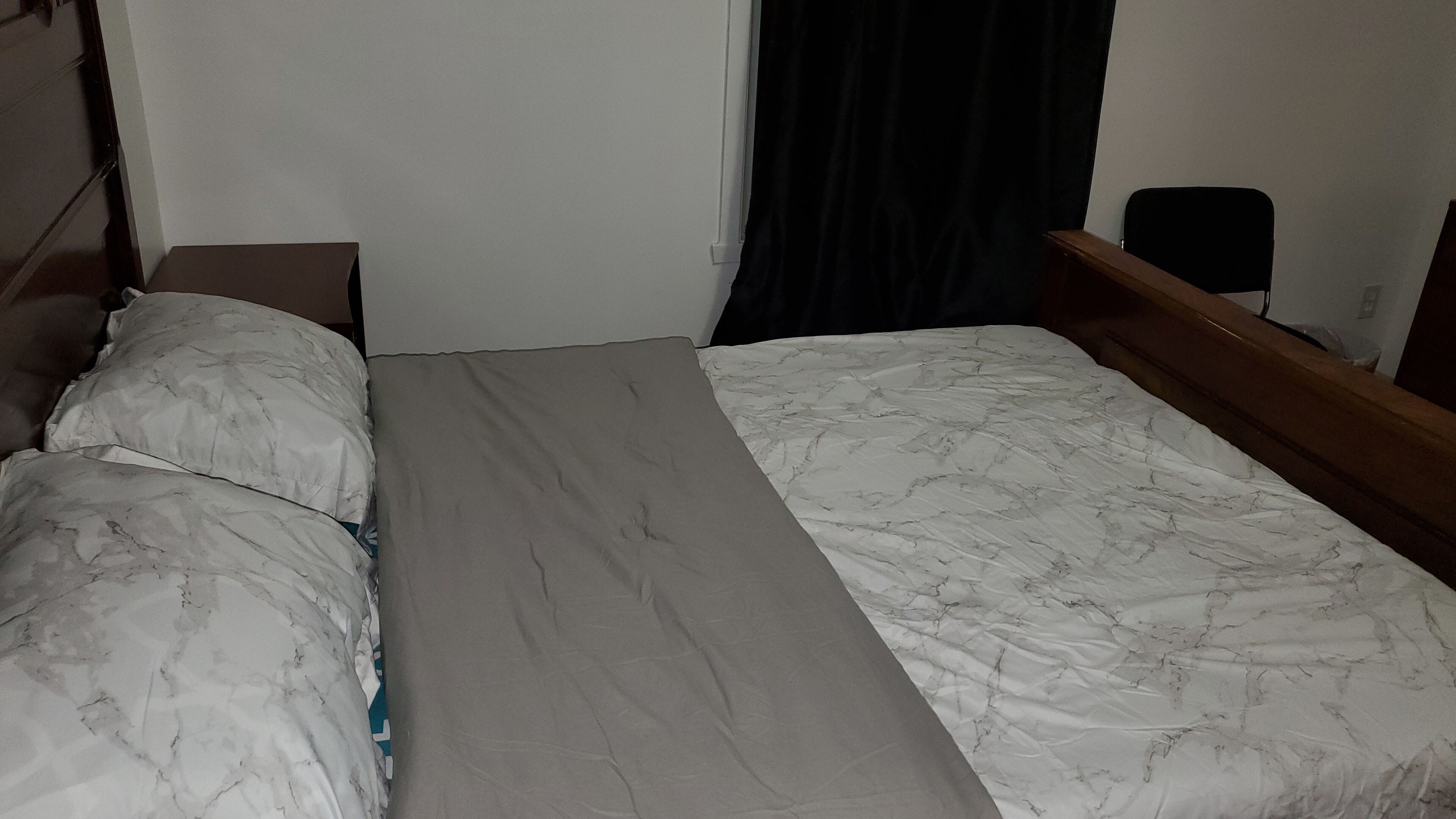 Una cama individual con colcha gris y blanca estampada, cabecera de madera y mesita de noche con lámpara.