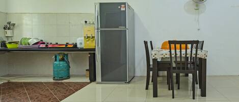 A kitchen with a refrigerator, a gas cylinder, a table with chairs, and a wall-mounted fan.