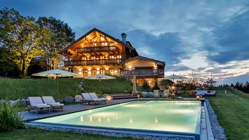 Ein großes, zweistöckiges Holzhaus mit Swimmingpool, Sitzgelegenheiten im Freien und einem gut beleuchteten Patio.