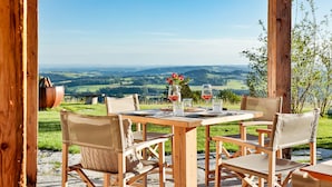 An outdoor dining area with a wooden table and chairs, a view of rolling hills, and a rustic wooden structure. - Alpenglühen-chalet-suite 90m², 2 Schlafzimmer, Wohnraum mit Küche und Bad mit Sauna (Frasdorf)