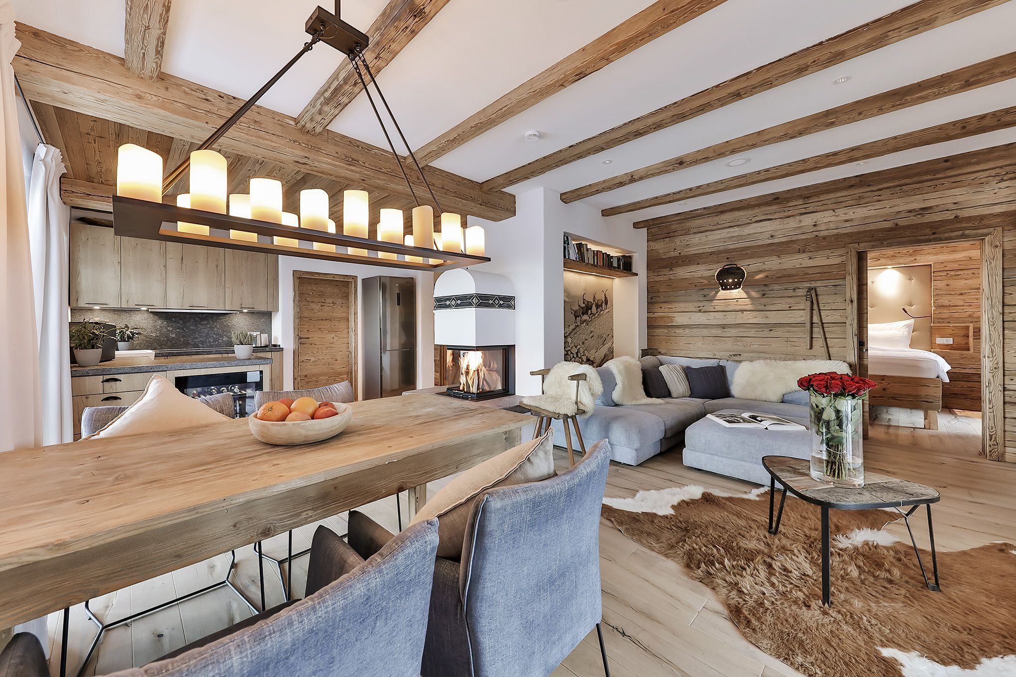 Ein modernes Interieur mit freiliegenden Holzbalken, einer Essecke mit Holztisch und -stühlen, einer Couch, einem kleinen Beistelltisch mit Blumenvase und einem Kamin.