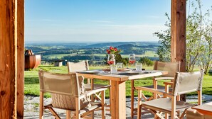 An outdoor dining area with a wooden table and chairs, a view of rolling hills, and a rustic wooden structure. - Gipfelglück-chalet-suite mit 60qm, Schlafzimmer, Wohnraum mit Küche und Bad mit Sauna (Frasdorf)