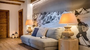 A cozy living room with a wooden floor, a grey sofa, two wooden nightstands with lamps, and a large mountain mural on the wall. - Gipfelglück-chalet-suite mit 60qm, Schlafzimmer, Wohnraum mit Küche und Bad mit Sauna (Frasdorf)