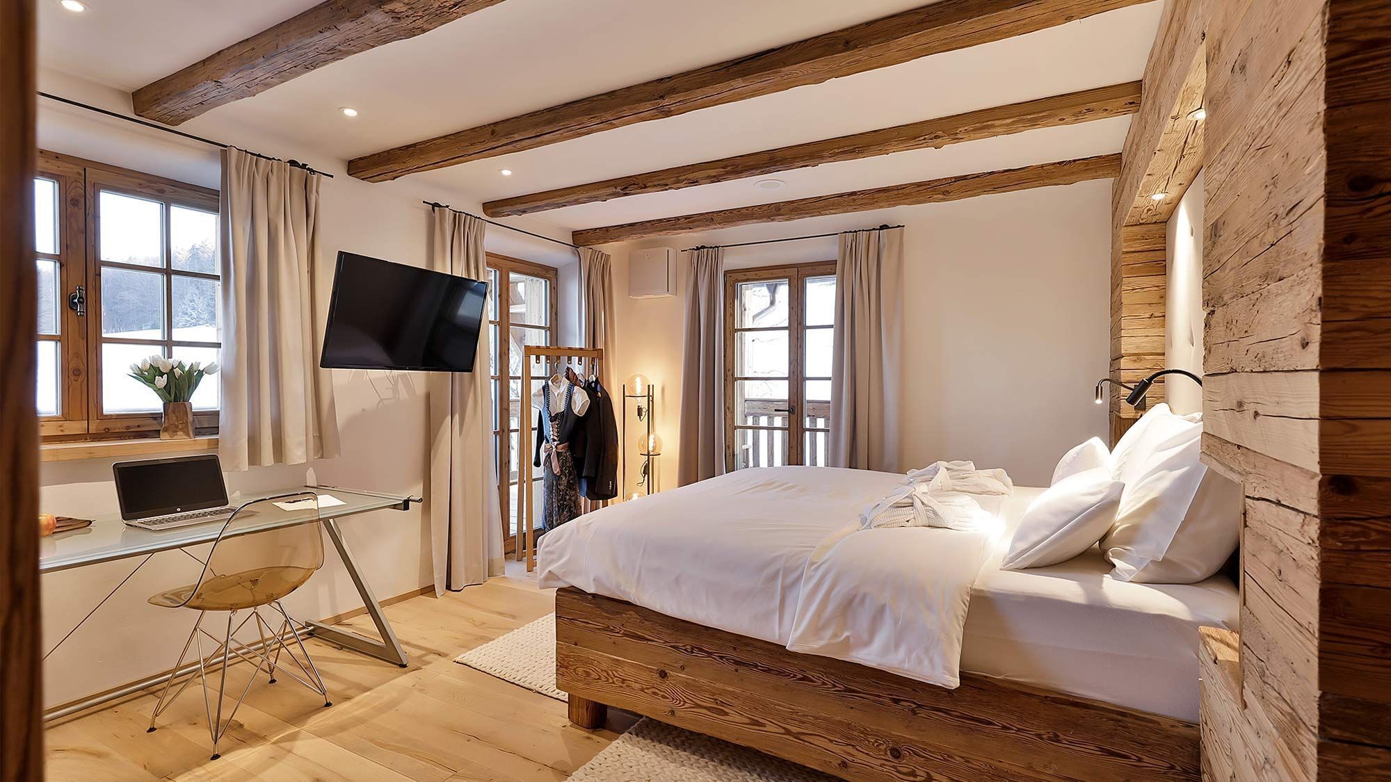 Ein Schlafzimmer mit einem hölzernen Bettgestell, einem Schreibtisch mit einem Laptop, einem Fernseher und einem Fenster mit Vorhängen.
