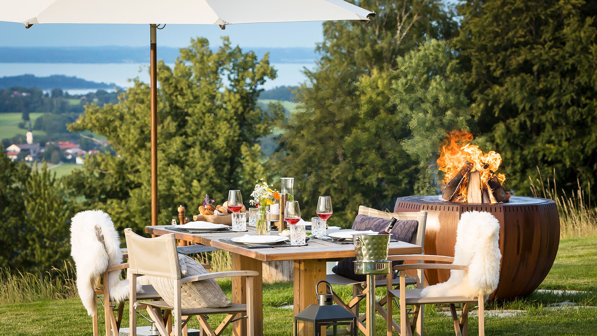 Un espace extérieur aménagé pour les repas, avec une table dressée, entourée de chaises, et un grand feu de camp en arrière-plan.