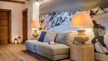 Ein gemütliches Wohnzimmer mit Holzboden, einem grauen Sofa, zwei Nachttischen aus Holz mit Lampen und einem großen Wandgemälde von Bergen.