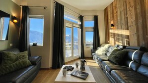 En stue med to lædersofaer, et sofabord med en bog og kop, samt store vinduer med gardiner.