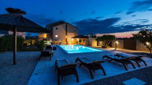 Una piscina con lettini e ombrelloni, circondata da un patio in pietra e un edificio con ingresso illuminato.