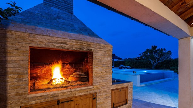 Camino esterno in pietra con fuoco acceso, inserito in una zona patio con piscina sullo sfondo.