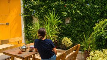 Una persona sentada a una mesa de madera con una tetera y una taza, en un jardín con una puerta amarilla y follaje verde.