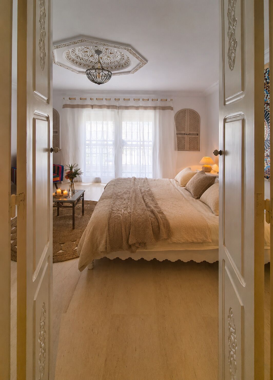 A bedroom with a bed, a chandelier, a table with candles, and a view of the outside through a window.
