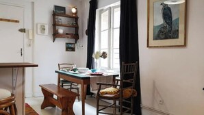 Un rincón comedor acogedor con una mesa y sillas de madera, un banco, una estantería con libros, un cuadro enmarcado en la pared y una ventana con cortinas.