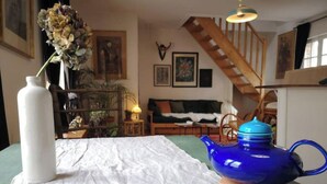Una habitación con una cama, una tetera azul, un jarrón blanco y un cuenco sobre una mesa. Se ve una escalera al fondo.