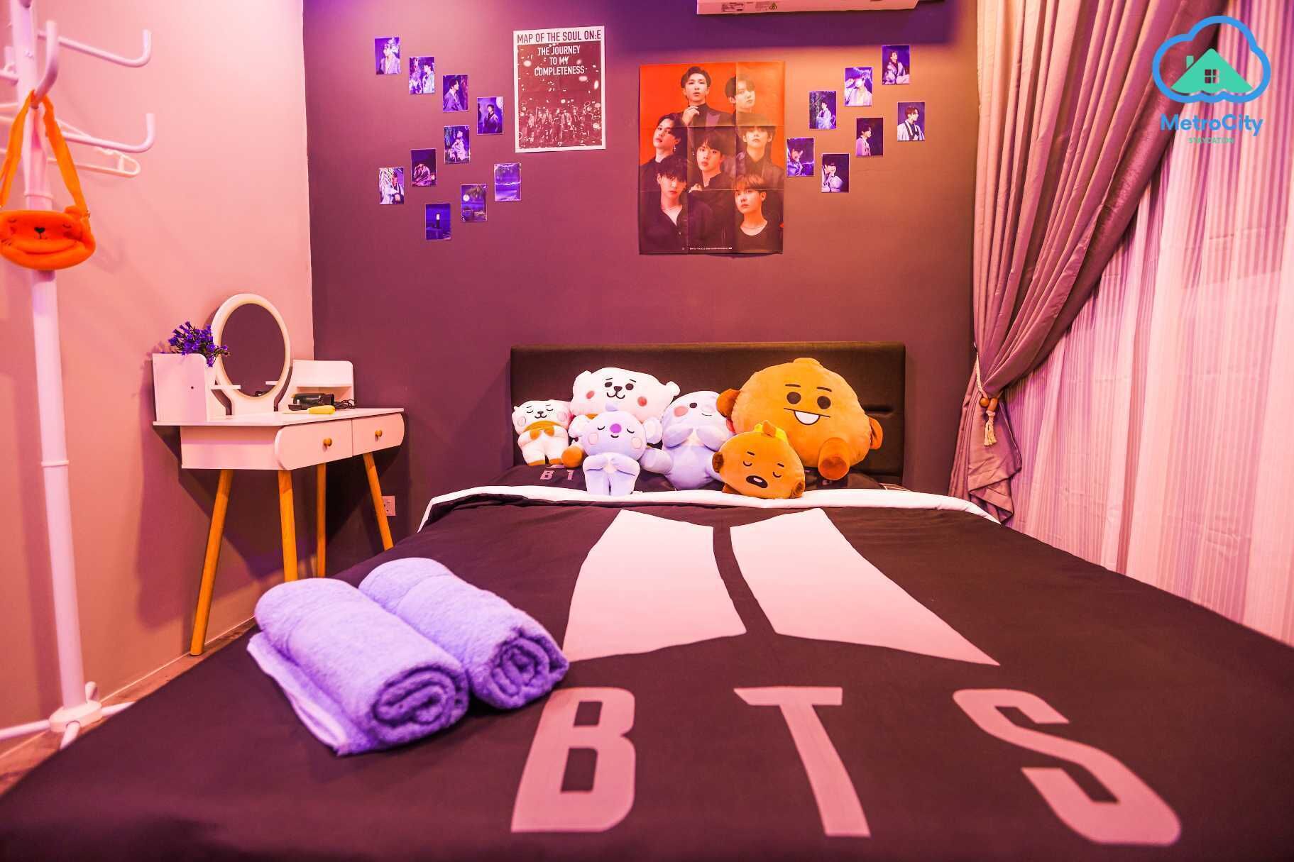 Une chambre avec un lit orné du logo de BTS, une commode avec un miroir, une petite table avec une plante et un mur décoré de photos et d’affiches.