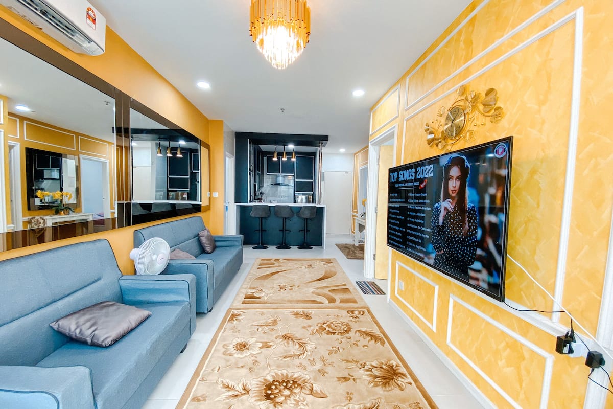 Una sala de estar moderna con un sofá, un televisor de pantalla plana grande mostrando 'TOP SONGS 2022', una cocina al fondo y un panel decorativo en la pared.