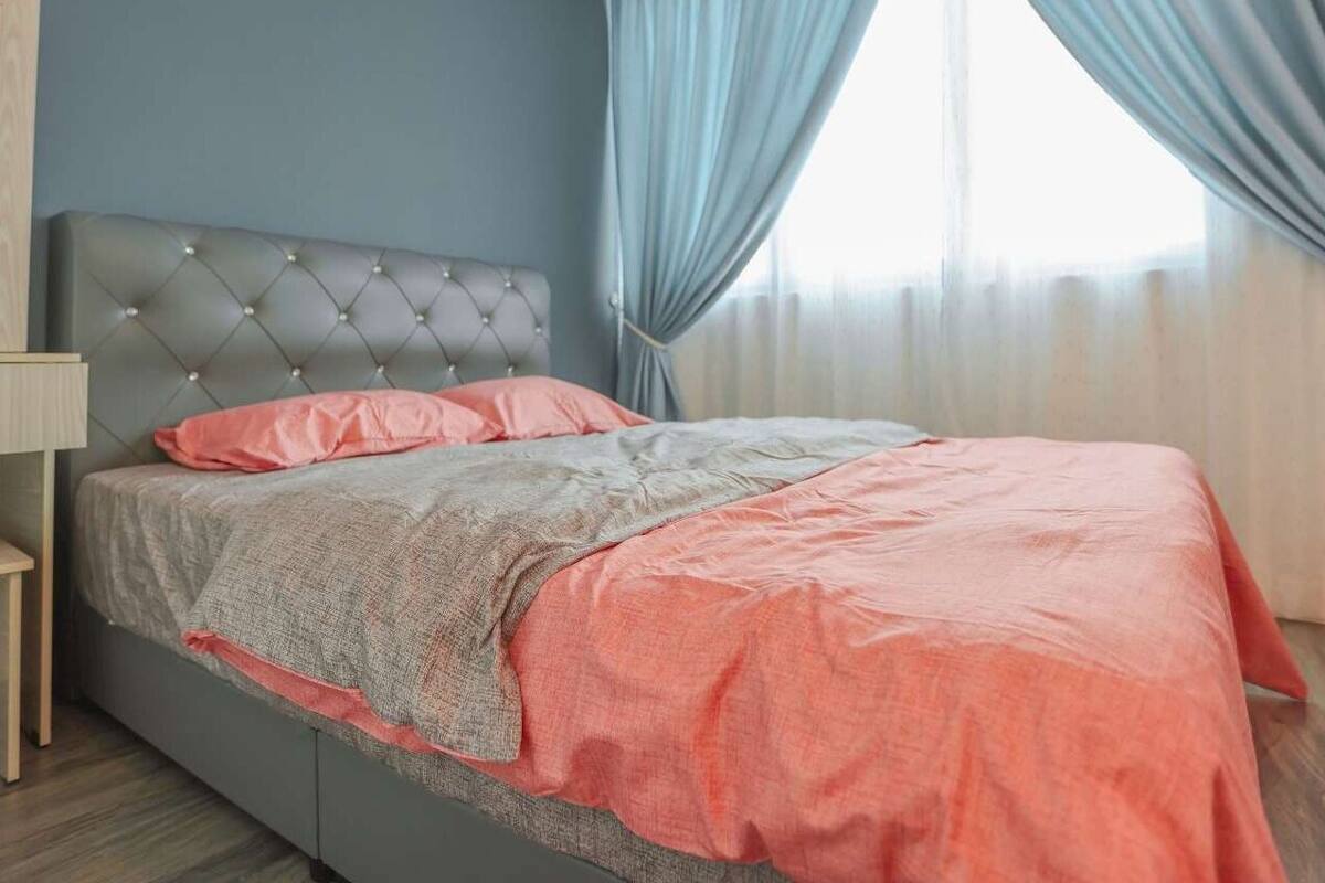Un dormitorio con cabecera capitonada, cama con ropa de cama rosa y gris, una mesita de noche y una ventana con cortinas.