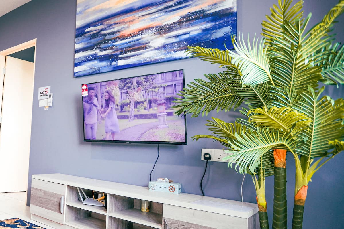 Una sala de estar con una televisión de pantalla plana montada en la pared, un mueble blanco para entretenimiento debajo, un tapete azul estampado y una palmera artificial de gran tamaño.