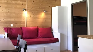 A compact room with a wooden panel wall, a white sofa with red cushions, a white cabinet, and a black refrigerator.