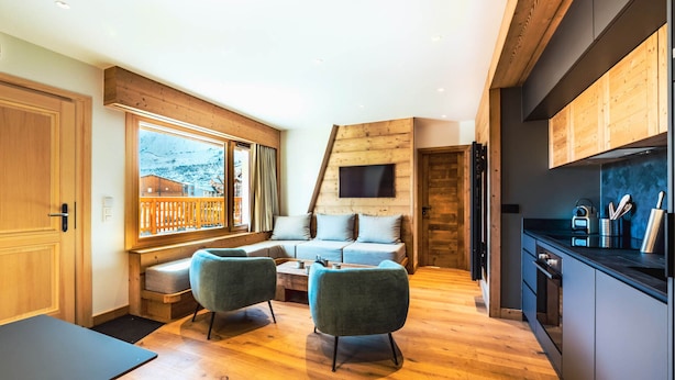Un interior moderno con suelo de madera, un sofá, dos sillones, una mesa de comedor y una zona de cocina con encimera y armarios negros.