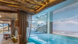 A modern spa room with a glass infinity pool, wooden ceiling, and a view of snowy mountains.