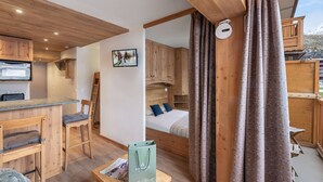 Ein gemütliches Zimmer mit Holzmöbeln, einem Bett mit blauen Kissen, einem Fernseher und einem Fenster mit Vorhängen.
