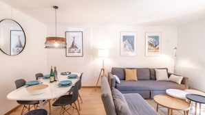 A modern living room with a grey sofa, a dining table with chairs, and a small coffee table. There are framed pictures on the wall and a lamp on the floor.