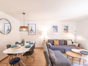 A modern living room with a grey sofa, a dining table with chairs, and a small coffee table. There are framed pictures on the wall and a lamp on the floor.