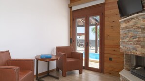Ein Zimmer mit Holzbalkendecke, Kamin aus Stein, Wandfernseher und Blick auf einen Swimmingpool durch eine Glastür.