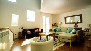 A spacious living room with a high ceiling, a large window, a radiator, a sofa, a coffee table, and a mirror on the wall. - Casa Boutique en Puerto Pesquero de Asturias (Viavélez)
