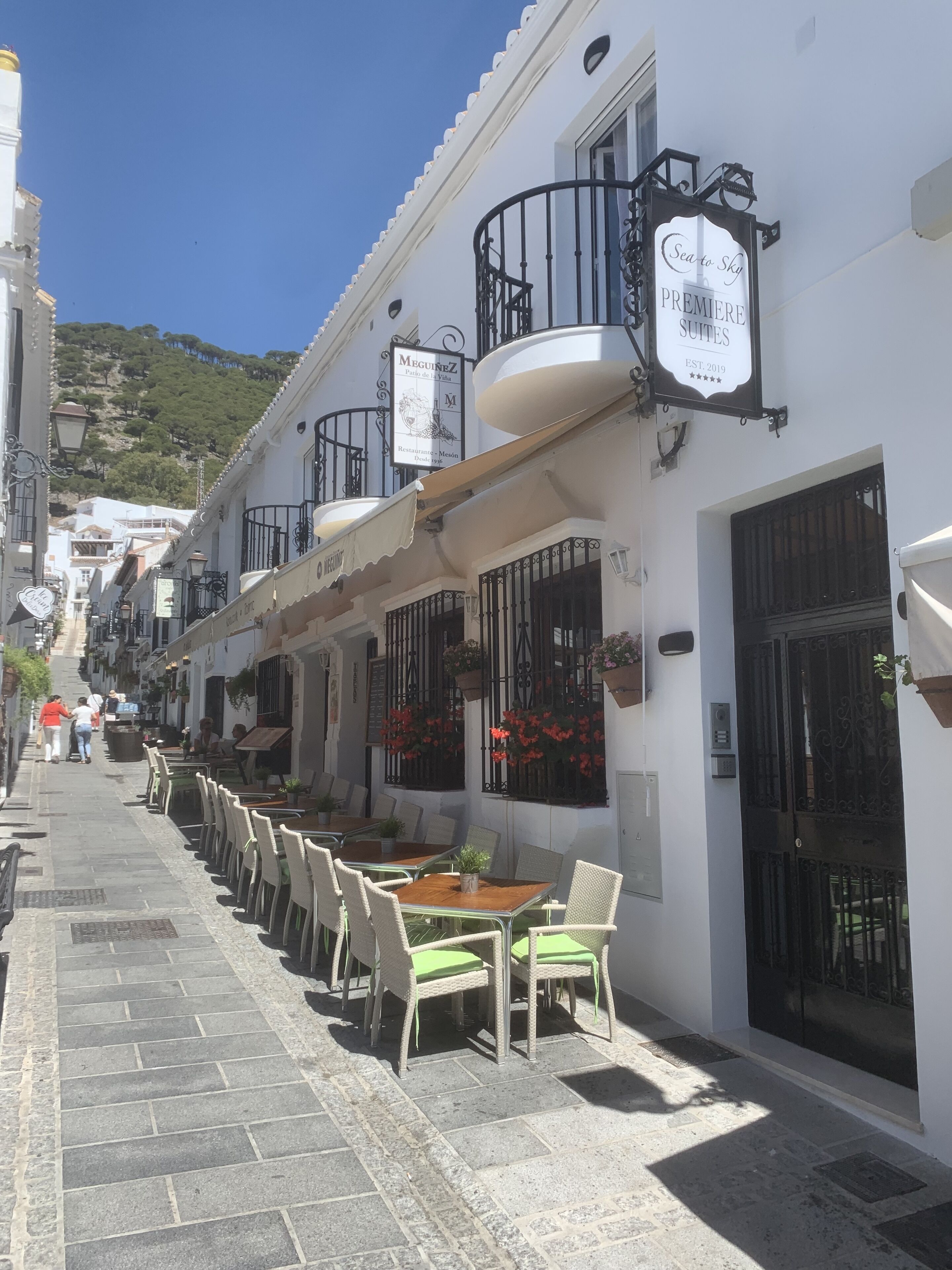 Una calle estrecha con mesas al aire libre, edificios blancos y cestas de flores colgantes.