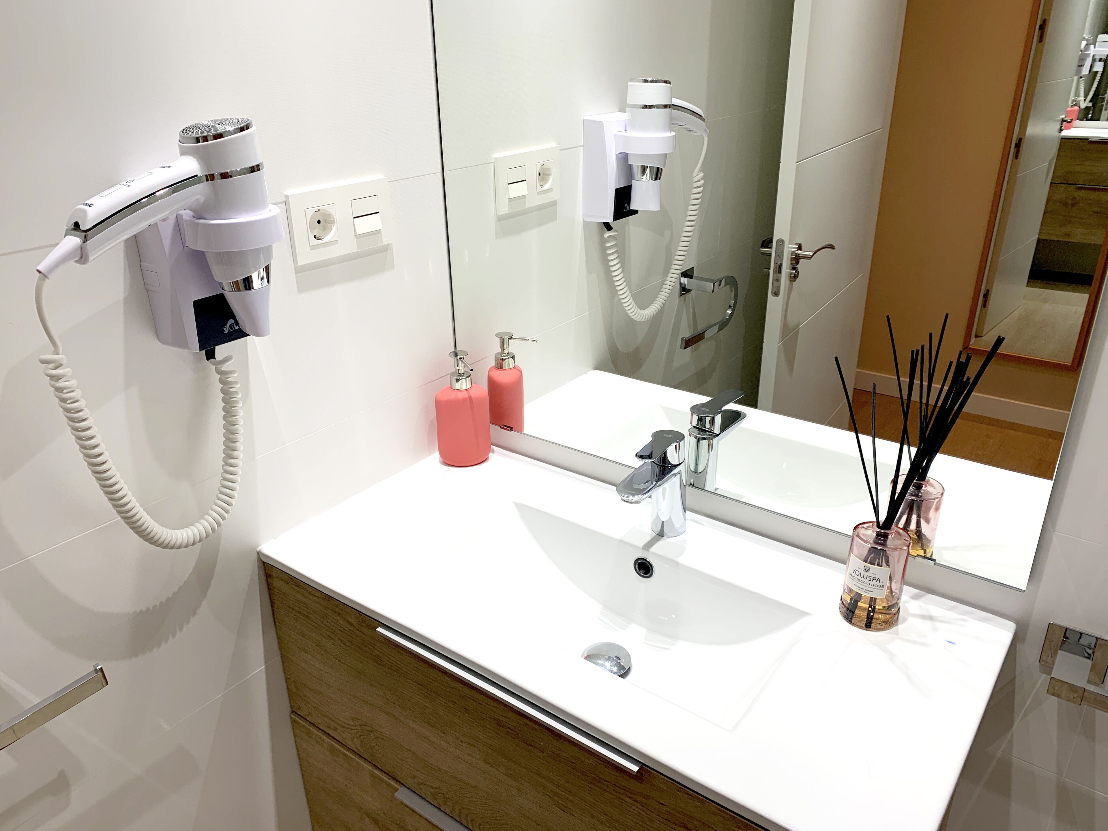 Un baño moderno con un lavamanos blanco, un espejo, un secador de pelo y una botella de champú.