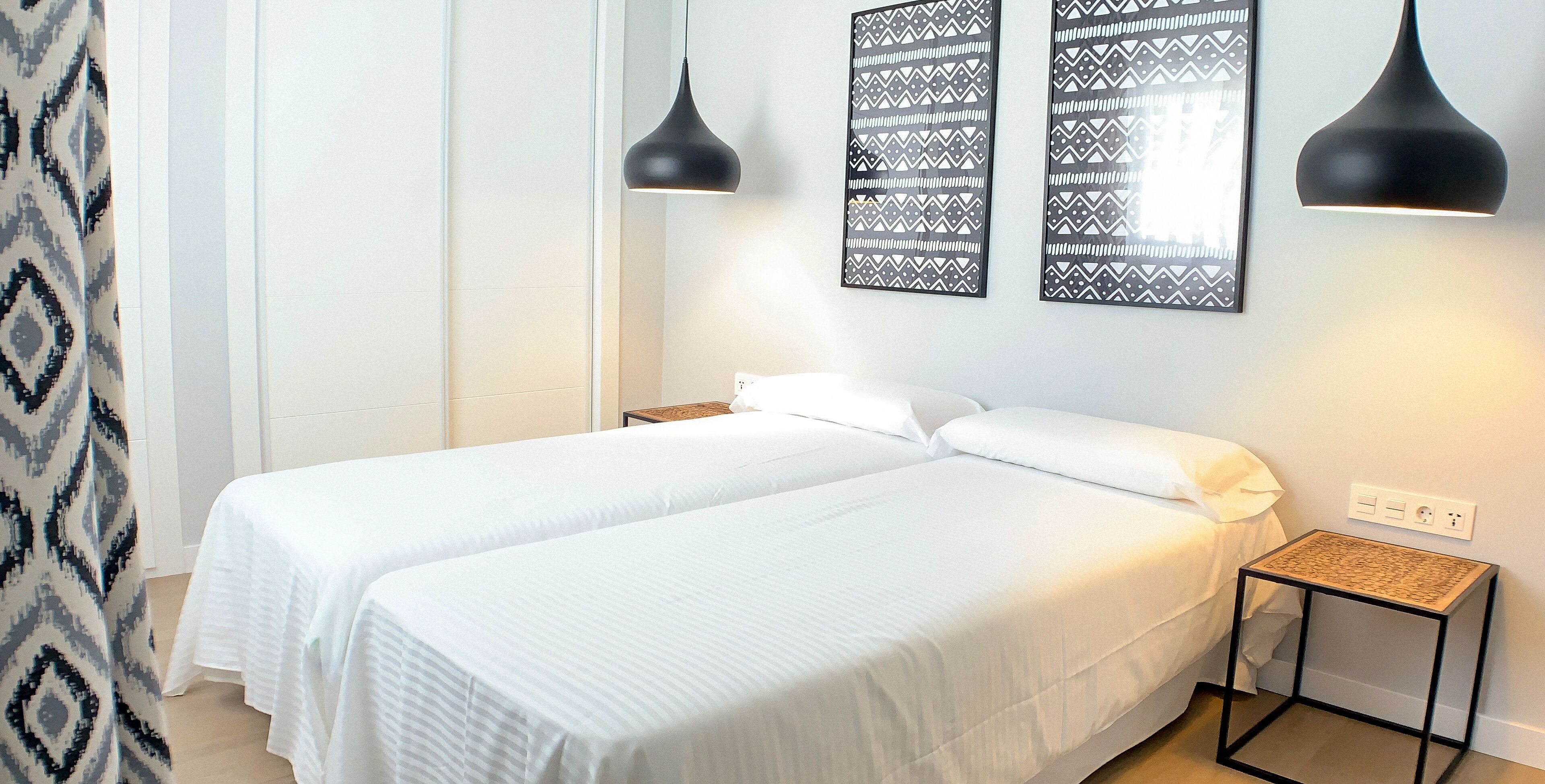 Habitación de hotel con dos camas individuales, una mesita de noche, una lámpara de pared y cortina estampada.