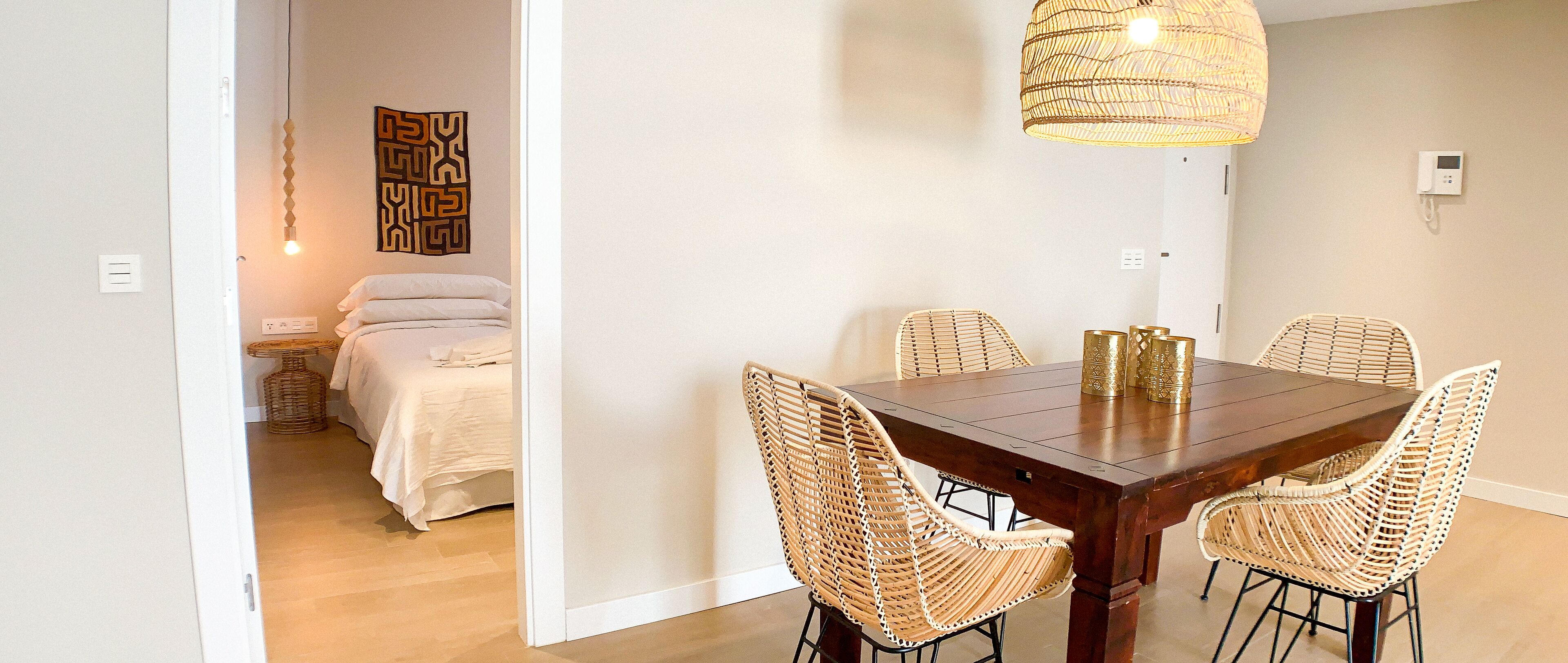 A dining area with a wooden table and wicker chairs, a hanging light fixture, and a bed with white linens in the background.