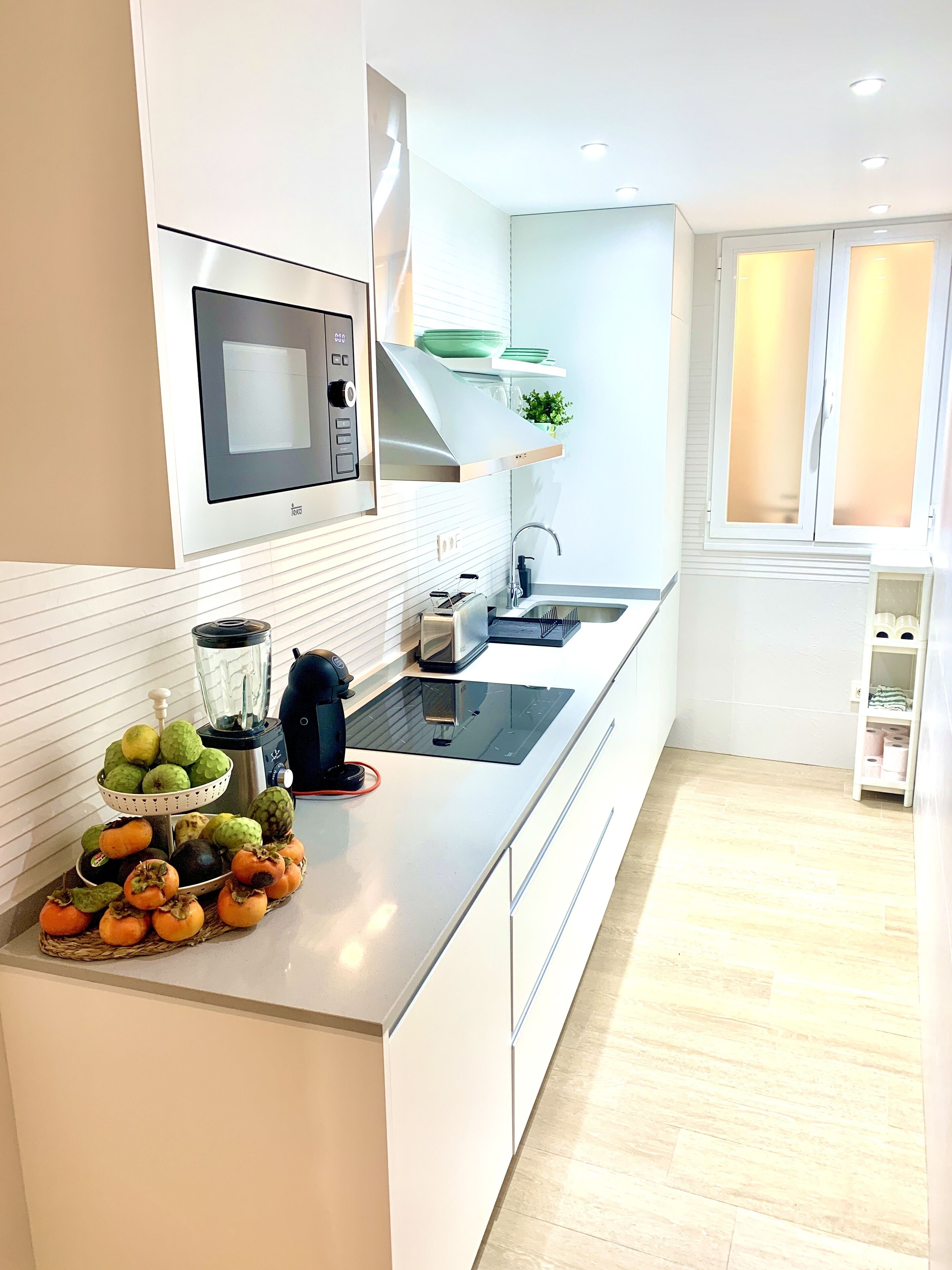 Una cucina moderna con mobili bianchi, forno a microonde integrato, cappa in acciaio inossidabile e un piano di lavoro con una ciotola di frutta e una macchina per il caffè.