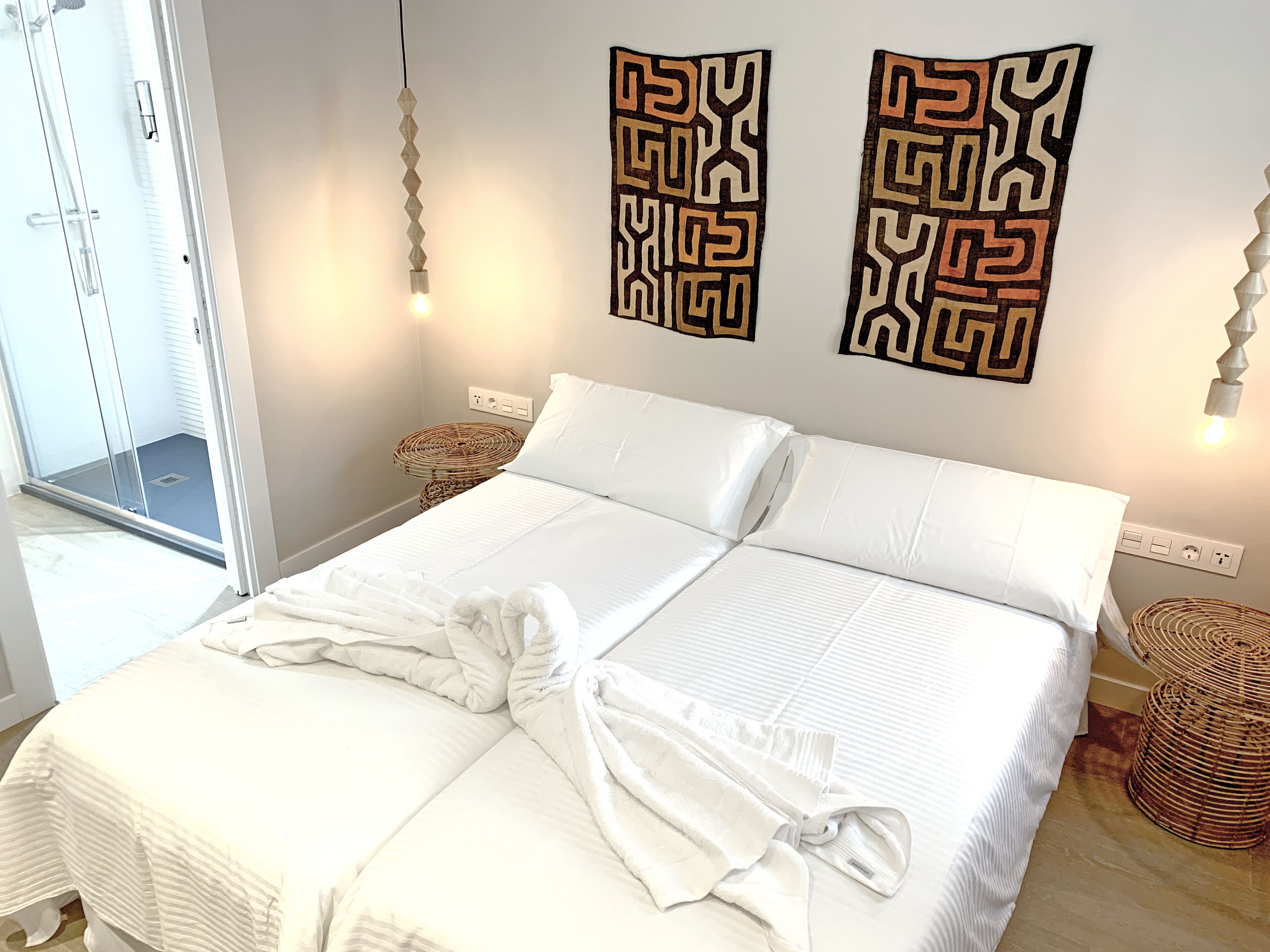 A double bed with white linens, two wall-mounted decorative panels, a bedside lamp, and a wicker side table.