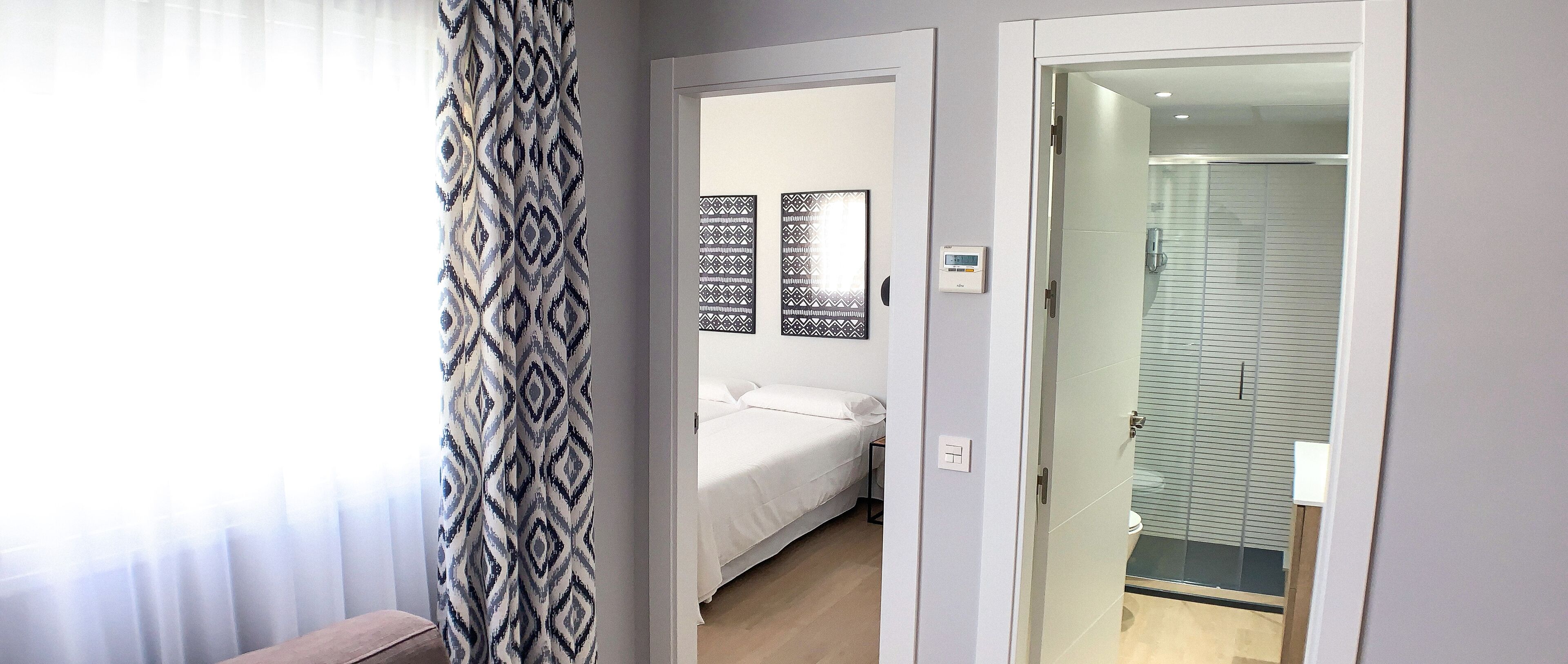 A bedroom with a bed, a bedside table, and a mirror. There is a bathroom with a shower and a toilet. The room has a modern design with light-colored walls and a patterned curtain.