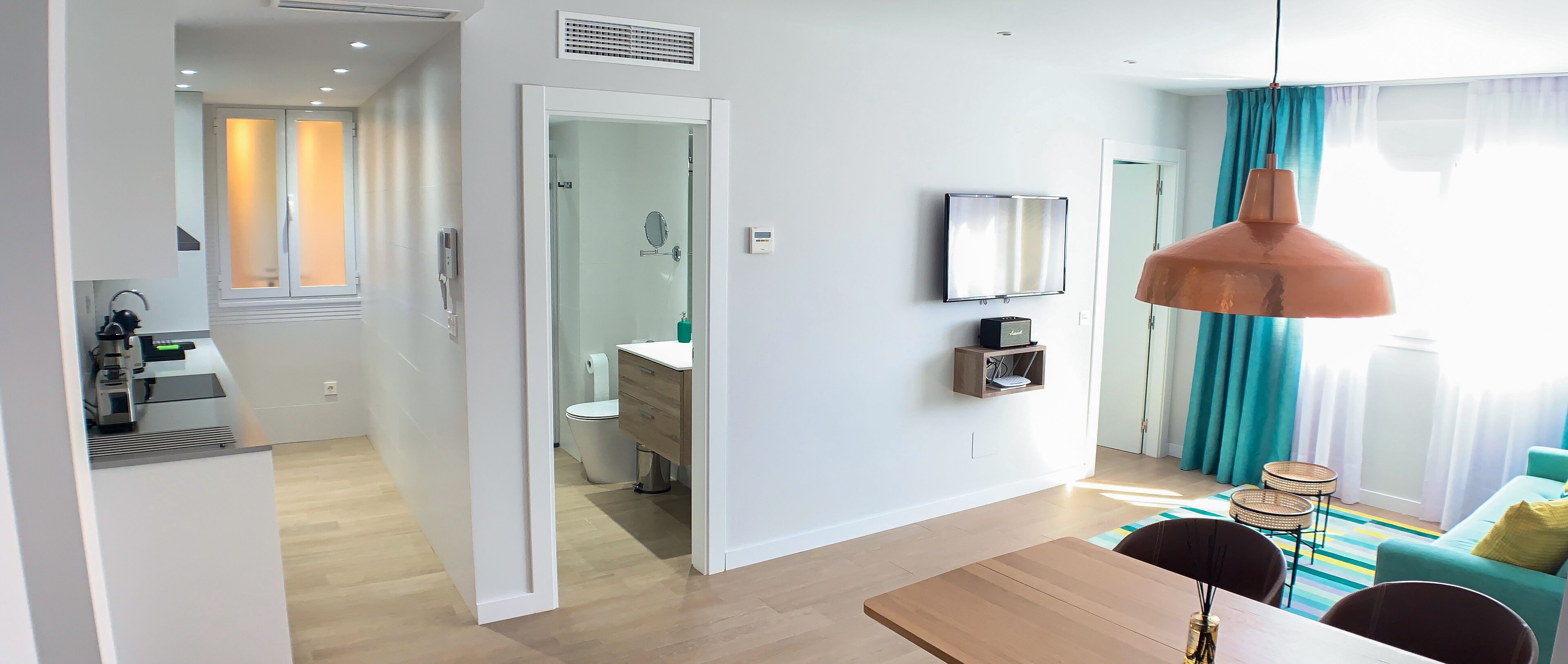 Habitación de hotel moderna con una mesa de comedor de madera, un espejo grande, una lámpara colgante y vistas al baño a través de una puerta abierta.