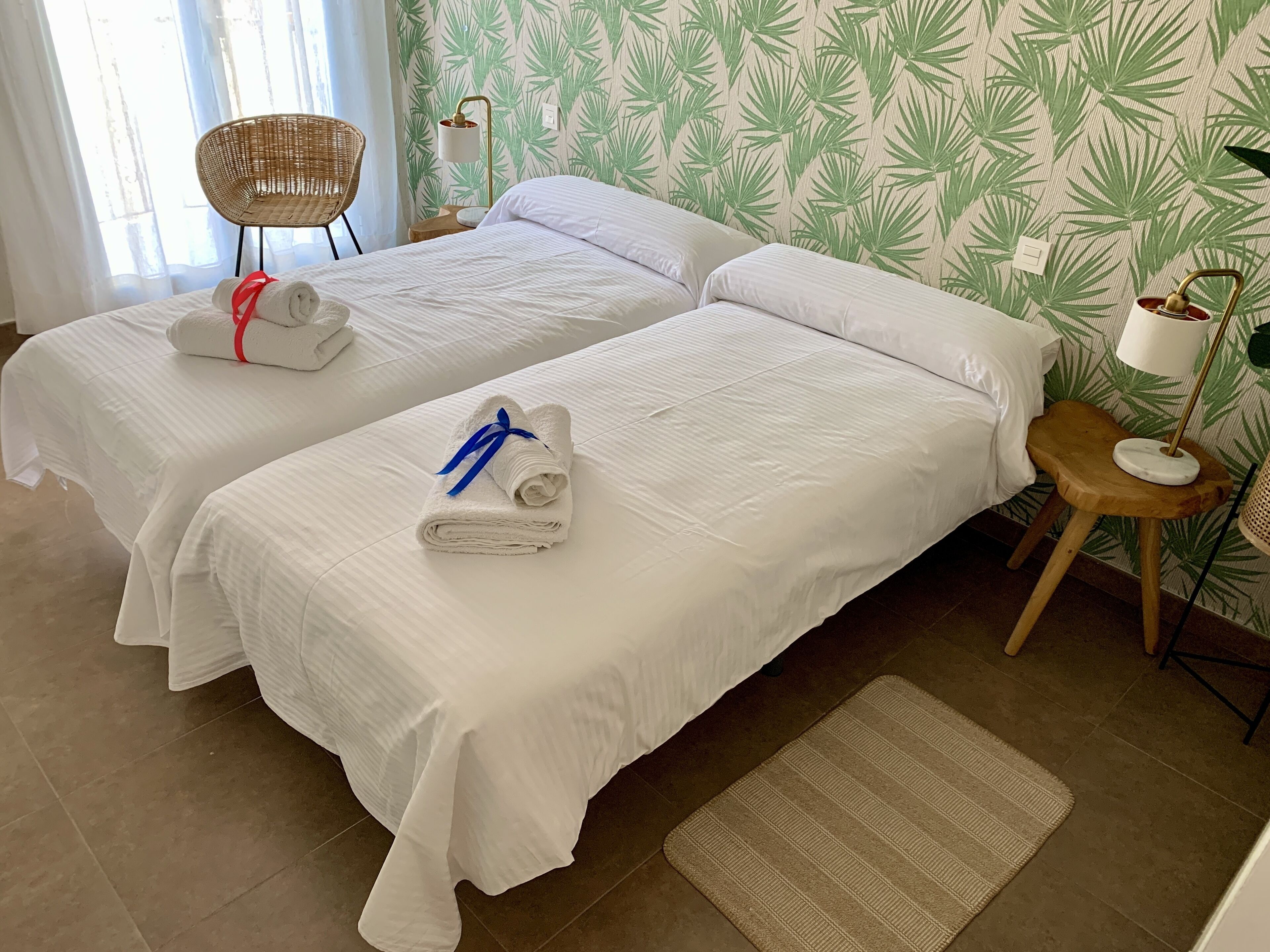 Camera d'albergo con due letti, una poltrona in vimini, un tavolino in legno, una lampada da terra e carta da parati decorata.