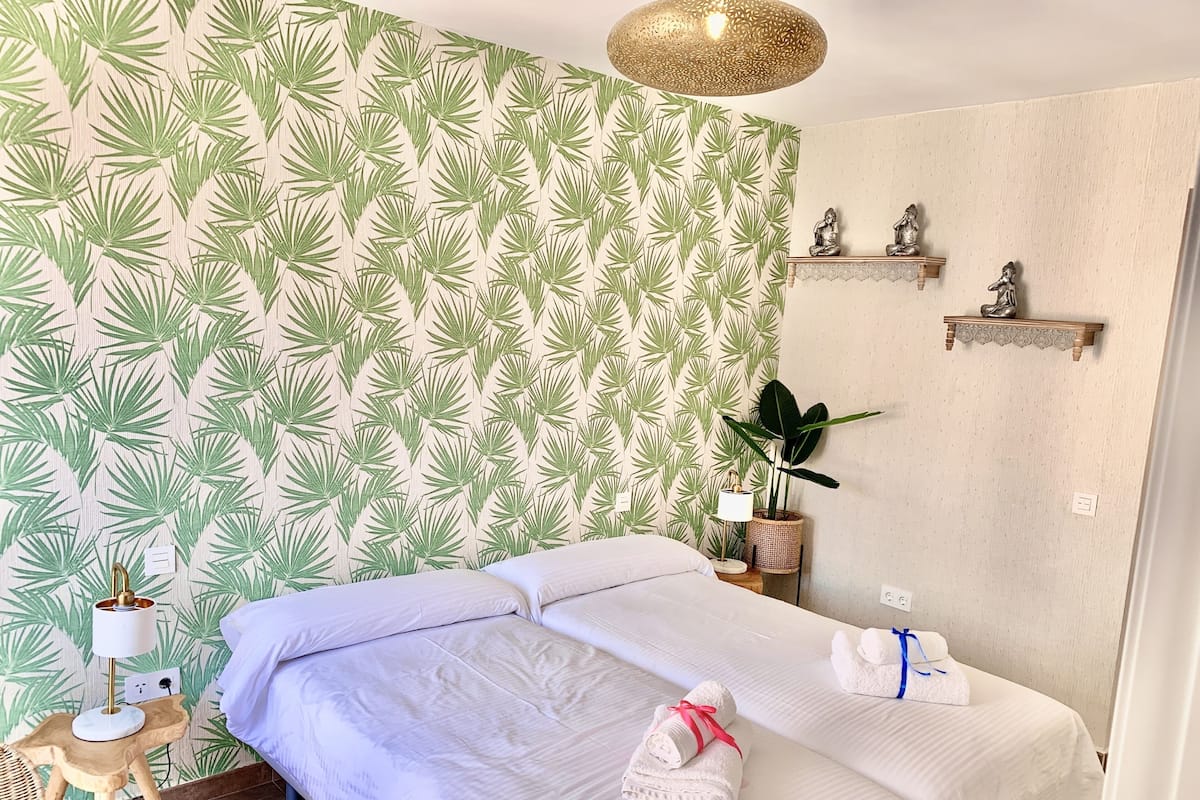 Habitació amb un llit, una taula petita amb una làmpada, una planta en test i prestatges fixats a la paret amb objectes decoratius.