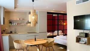 Een moderne hotelkamer met kitchenette, een eethoek met tafel en stoelen, een bed met rode accentmuur, en een aan de muur bevestigde televisie.