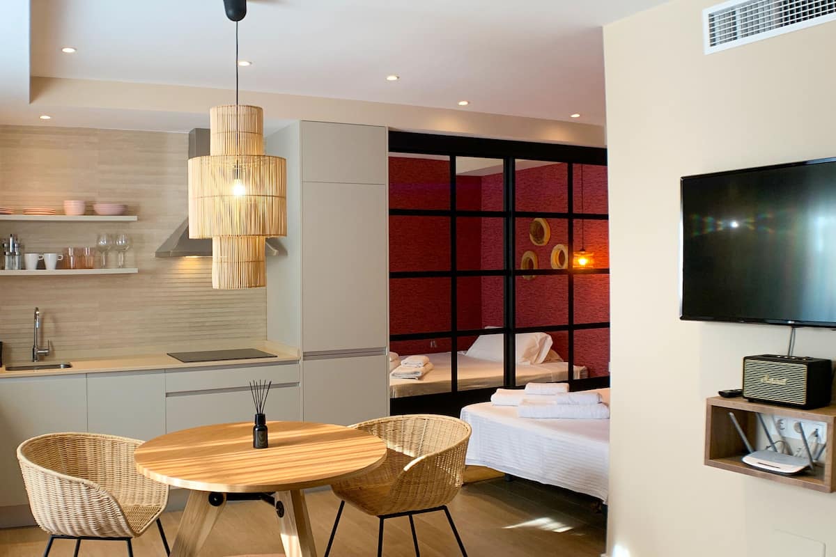 Habitació d'hotel moderna amb cuineta, zona de menjador amb taula i cadires, llit amb paret decorativa vermella i televisió penjada a la paret.