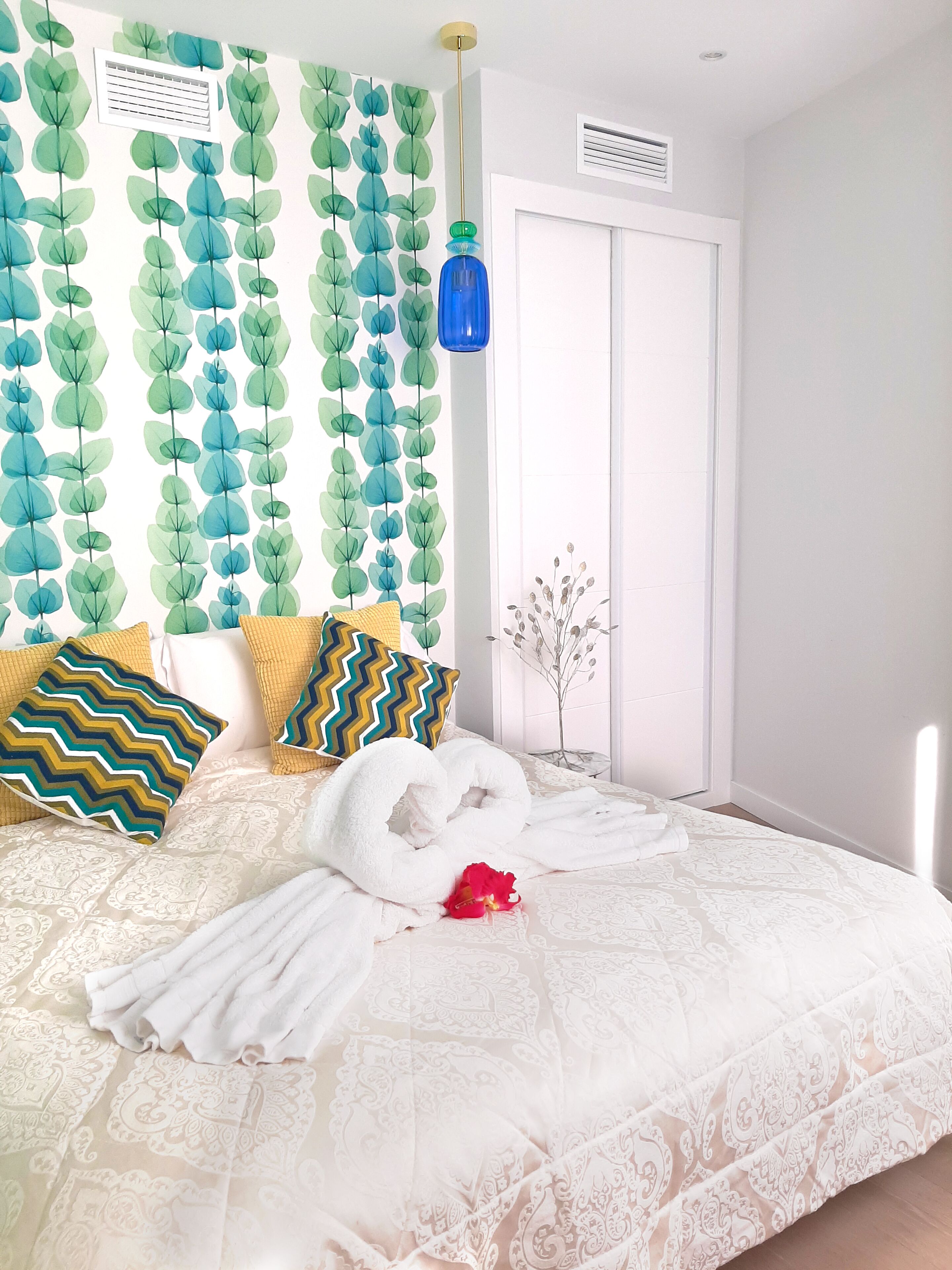 Un dormitorio con una cama, almohadas, una pared con diseño de hojas, una lámpara colgante azul y una puerta.